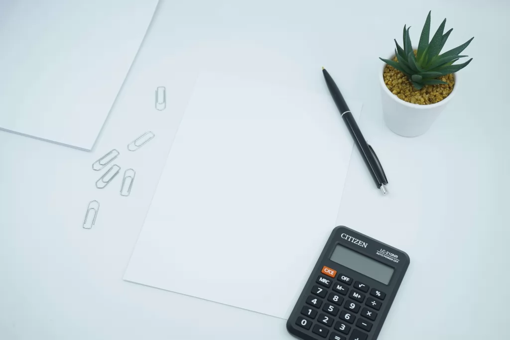 kalkulator, długopis, spinacze, kartki papieru i mała roślina doniczkowa na białym biurku - polsko niemieckie biuro rachunkowe